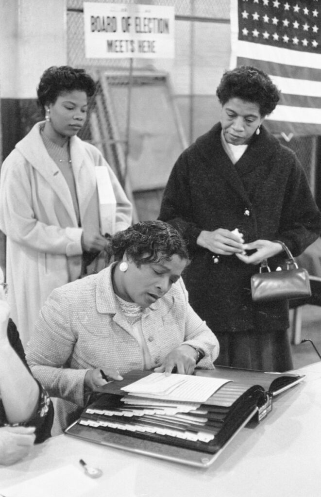 Three African American women at a polling place, one looking at a book of registered voters on November 5, 1957, in New York City or Newark, New Jersey.